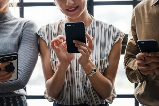 Three People Stood Next to Each Other Scrolling on Their Smartphones