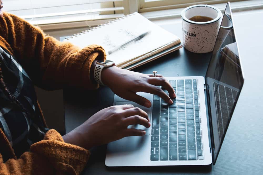 A person typing on a laptop with a planner and mug of tea in the background.