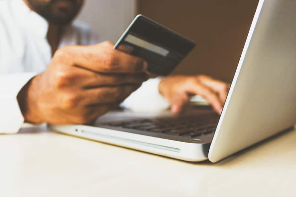 A man holds up a credit card while typing on his laptop with the other hand.